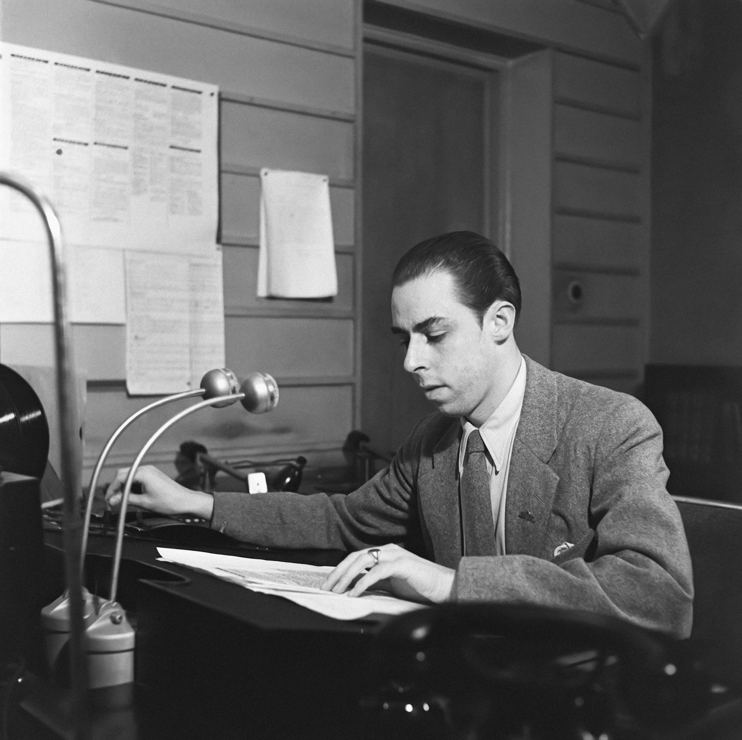 A man sitting at a desk in front of microphones, reading notes and turning a knob